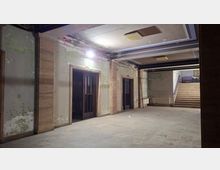 Verlassene Innenhalle eines älteren Gebäudes mit abblätternder Wandfarbe, Steinverkleidungen an Pfeilern und zwei dunklen Holztüren. Rechts führt eine breite Treppe nach oben; an der Decke sind rechteckige Leuchten und freiliegende Kabel zu sehen.