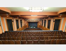 Innenansicht eines Theatersaals mit ansteigenden Sitzreihen, einer zentralen Bühne mit schwarzem Vorhang und einer Beleuchtungsanlage an der Decke. Die Wände sind orangefarben mit akustischen Elementen, und die Bühne ist von hölzernen Verkleidungen eingerahmt.