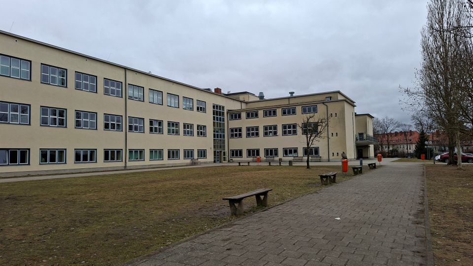 Großer Innenhof vor einem mehrstöckigen, hellbeigen Schul- oder Verwaltungsgebäude mit vielen rechteckigen Fenstern und einem verglasten Treppenhaus. Daneben verläuft ein gepflasterter Weg mit Bänken und einzelnen Bäumen auf einer Rasenfläche unter bewölktem Himmel.