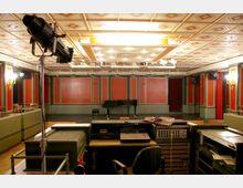 Ein kleiner Konzert- oder Veranstaltungssaal mit einem Flügel auf der Bühne, umgeben von rot und grün gestrichenen Wänden mit goldenen Verzierungen sowie Holzelementen. Im Vordergrund ist ein technischer Steuerplatz zu sehen, einschließlich einer Mischkonsole und Beleuchtungsequipment.