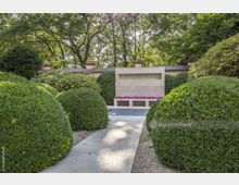 Ein gepflegter Garten mit rund geschnittenen Buchsbaumsträuchern, einem gepflasterten Weg und einer modernen Sitzbank mit pinkfarbenen Kissen vor einer niedrigen, beige Steinmauer. Im Hintergrund sind hohe Bäume und ein Zaun mit Ziegelpfeilern zu sehen.
