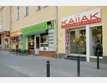 Straßenansicht mit mehreren Geschäften in einem gelben Gebäude; links ein Döner-Imbiss, in der Mitte ein Spätkauf mit grünem Schild und Eistheke, rechts ein Schmuckgeschäft mit Glastür und weiß-rotem Schild. Davor stehen wenige Außentische und vereinzelt Poller auf dem Gehweg.