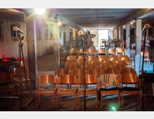 Ein einfacher Veranstaltungsraum in einem alten, teilweise verfallenen Gebäude mit Reihen aus Holzstühlen, Mikrofonständern im Vordergrund und sichtbaren, unverputzten Betonsäulen. Im Hintergrund befindet sich diverses gestapeltes Mobiliar, und Tageslicht fällt durch eine offene Tür.