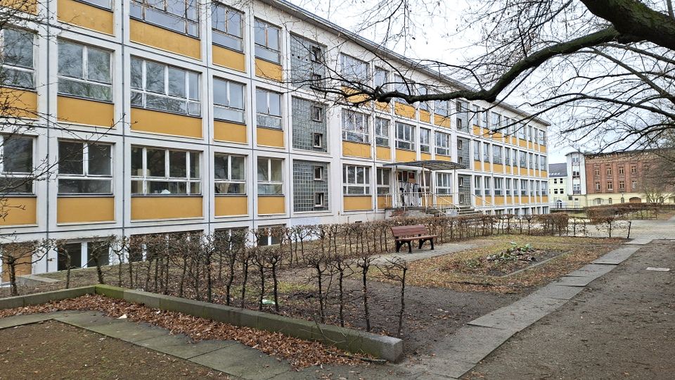 Mehrstöckiges Schul- oder Verwaltungsgebäude mit gelb-weißer Rasterfassade und vielen rechteckigen Fenstern, davor ein kleiner Hof mit Hecken, Wegen und einer Bank. Kahle Baumäste ragen über den Vorplatz, im Hintergrund sind weitere Gebäude zu sehen.