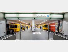U-Bahn-Station mit gelben Zügen der Linie U7 auf beiden Gleisen, umgeben von hell beleuchteten Bahnsteigen. Über dem Zugang hängt ein Schild mit der Aufschrift "Rathaus Spandau U7", im Hintergrund sind orangefarbene Wände mit Informationsplakaten zu erkennen.