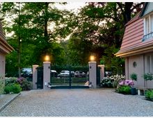 Einfahrt mit gepflastertem Weg, flankiert von zwei Häusern mit roten Ziegeldächern. Im Hintergrund befindet sich ein metallisches Tor mit Laternen auf gemauerten Pfeilern, dahinter parken Autos unter hohen Bäumen.