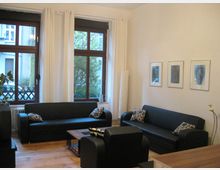 Wohnzimmer mit zwei schwarzen Sofas und einem schwarzen Sessel, einem kleinen Holztisch und dekorativen Kissen. Große Fenster mit weißen Vorhängen lassen Tageslicht herein, während die Wände mit drei gerahmten Bildern dekoriert sind.