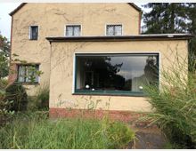 Gelbes Einfamilienhaus mit Putzfassade, teilweise bewachsen mit Pflanzen, und einem großen Fenster im Erdgeschoss. Im Vordergrund befindet sich ein verwilderter Garten mit hohen Gräsern und Büschen.