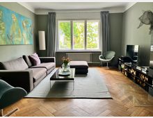 Wohnzimmer mit holzfarbenem Fischgrät-Parkettboden, modernen grauen Wänden und großen Fenstern mit grauen Vorhängen. Eine graue Eckcouch mit lila Kissen, ein Glastisch auf einem grauen Teppich, und ein Fernseher auf einem schwarzen Lowboard prägen den Raum. An der Wand hängt ein großes Gemälde in Blau- und Grüntönen.