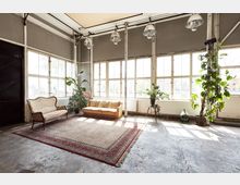 Industrie-Loft mit großen Fenstern und Betonboden, eingerichtet mit einem gemusterten Teppich, zwei Sofas im Vintage-Stil und mehreren großen Topfpflanzen. Die hohen Decken sind mit Industrielampen ausgestattet, und die Wände zeigen freiliegende Stahlträger und teilweise Ziegel.