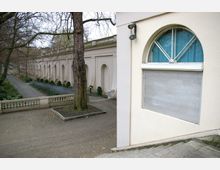Ein Gebäude mit einer hellen Fassade und einem halbkreisförmigen Fenster mit blauer Gardine im Vordergrund, daneben eine Baumreihe entlang eines schmalen Gehwegs und eine Balustrade, die das Gelände abgrenzt. Im Hintergrund verläuft eine arkadenartige Mauer mit Rundbögen, umgeben von Pflanzen.