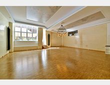 Großer, heller Raum mit Holzfußboden und cremefarbenen Fliesenwänden. Eine große Fensterfront lässt Tageslicht herein, während eine moderne Deckenleuchte und ein verziertes Deckenelement das Innere schmücken; im Hintergrund sind kleine Treppenstufen sichtbar.