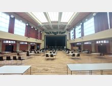 Großer Veranstaltungsraum mit Parkettboden, mehreren rechteckigen Tischen und Stühlen, einer erhöhten Bühne mit Vorhängen und Scheinwerfern sowie hohen Fenstern, die von roten Vorhängen flankiert werden. Die Decke hat ein großes Oberlicht, das für viel Tageslicht sorgt.