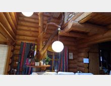 Innenraum eines rustikalen Blockhauses mit sichtbaren Holzstämmen an Wänden und Decke. Eine runde, hängende Lampe leuchtet über einem dekorierten Regal mit Pflanzen und Porzellan; bunte Vorhänge und ein großes Holzfenster ergänzen die Einrichtung.