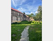 Innenhof mit Rasenfläche und schmalem gepflastertem Weg, umgeben von alten Backsteinmauern mit zugemauerten Bögen und einem Gebäude mit rotem Dach. Auf der Wiese stehen mehrere einfache Holzbänke und Tische, daneben Bäume und Sträucher am Rand.