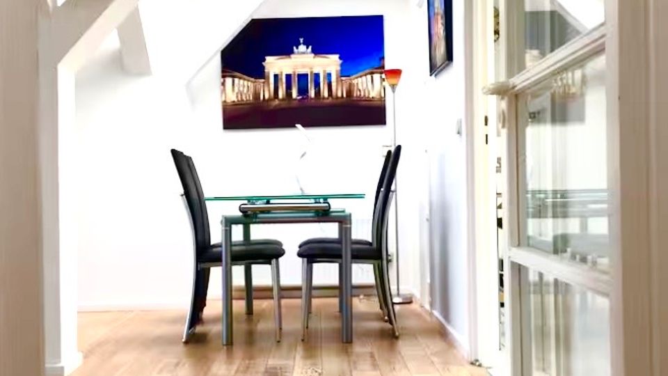 Heller, schmaler Innenraum mit sichtbaren weißen Dachbalken und hellem Holzboden, der zu einer kleinen Essecke führt. Am Ende steht ein Glastisch mit zwei schwarzen Stühlen, darüber hängt ein Wandbild; rechts sind große verglaste Tür- oder Fensterrahmen.