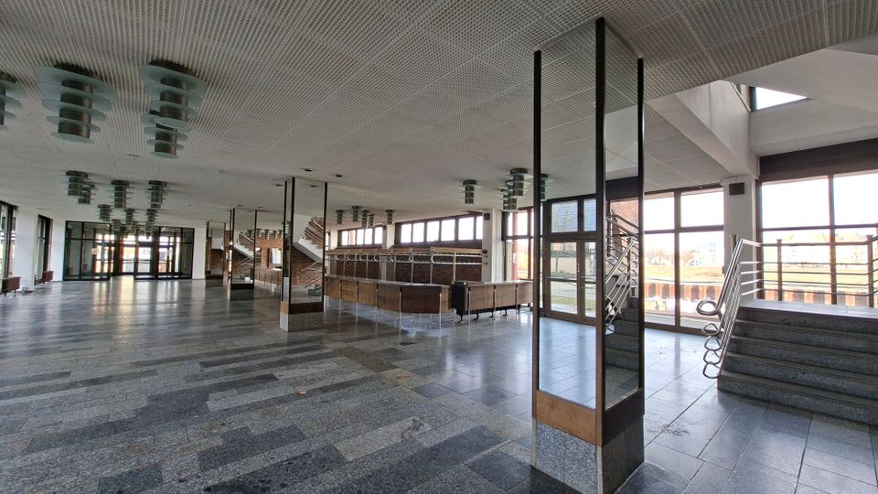 Weitläufige Eingangshalle eines öffentlichen Gebäudes mit grauem Steinboden, abgehängter Decke mit runden Leuchten und großen Fensterfronten. Rechts führen breite Treppen mit Metallgeländern zu einem erhöhten Bereich; im Hintergrund sind Glastüren und ein Kassen- oder Tresenbereich sichtbar.