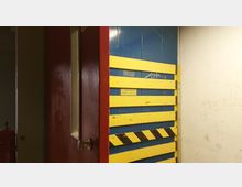 Ein geöffneter roter Aufzugsschacht mit einer gelben Sicherheitsbarriere aus horizontalen Streifen, die vor einer blauen Wand angebracht ist. Ein Hinweisschild zeigt die maximale Kapazität von 20 Personen oder 1500 kg an.