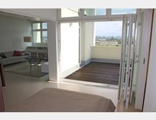 Wohnraum mit hellen Wänden, einem Sofa und Regalen im Hintergrund, verbunden mit einer offenen Holzterrasse durch eine große Glastür. Im Außenbereich ist ein Blick auf die Stadtlandschaft erkennbar.