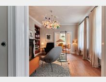 Wohnzimmer mit Holzboden und großen Fenstern mit bodenlangen Vorhängen. Möblierung umfasst ein graues und ein schwarzes Sessel, ein orangenes Sofa und eine bunte Hängelampe; im Hintergrund ein angrenzender Essbereich mit Holztisch und gelber Polsterstuhl.