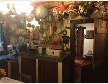 Gemütliche Landhausküche mit Holzwänden und -möbeln, dekoriert mit getrockneten Blumen, hängendem Kochgeschirr und antiken Haushaltsgegenständen. Im Vordergrund ein gedeckter Tisch und im Hintergrund eine geöffnete Tür, die in einen anderen Raum mit rotem Teppich führt.