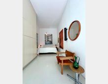 Narrow, minimalist bedroom with white walls and a glossy light floor, featuring a low bed with white bedding at the far end beneath a large framed artwork. Along the right wall are a wooden chair, small round side table with a green glass pitcher, a round wall mirror, and mounted wall art, while the left side has built-in white cabinetry.