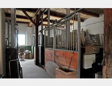 Innenansicht eines alten Stalls mit Holzbalkendecke, Metallgitter-Trennwänden und Holzelementen. Der Raum ist als Lager genutzt, gefüllt mit Möbeln, Kartons und sonstigen Gegenständen.