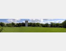 Weitläufige Parkanlage mit einer großen, gepflegten Rasenfläche, umgeben von Bäumen und Sträuchern; im Hintergrund ein modernes Beton-Denkmal mit zwei nach oben ragenden Elementen, dahinter niedrige Gebäude.