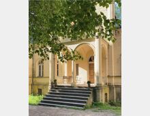 Sandfarbenes historisches Gebäude mit einer kleinen Treppe, die zu einer verandaartigen Eingangsfront mit drei großen Bögen führt. Umgeben von grünen Bäumen, sind auf der Veranda ein Holztisch und Stühle sowie dekorative Pflanzgefäße zu sehen.