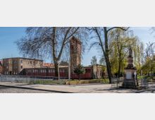 Historisches Feuerwehrgebäude mit rotem Tor und hohem Backsteinturm, im Vordergrund eine verzierte Steinfigur auf einem Sockel umgeben von Geländer, umgeben von Bäumen und einem gepflasterten Gehweg.