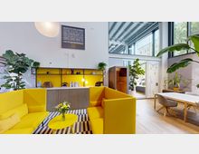 Modern eingerichteter Innenbereich mit gelben Polstermöbeln, einem kleinen Couchtisch mit Blumenstrauß und Wandregalen in Gelb und Schwarz. Große Fenster lassen viel Licht herein, während zahlreiche Zimmerpflanzen eine grüne, freundliche Atmosphäre schaffen.