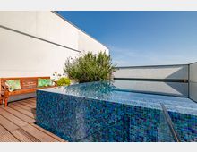 Rooftop-Terrasse mit einem rechteckigen Infinity-Pool, dessen Kacheln in verschiedenen Blautönen schimmern. Im Vordergrund befinden sich Holzdielen, eine Holzbank mit bunten Kissen und üppige Pflanzen neben einer weißen Wand.