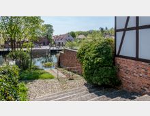 Ein gepflasterter Weg führt entlang einer Treppe zu einem kleinen Kanal, der von Bäumen und Sträuchern umgeben ist. Im Hintergrund sind eine Brücke, rote Backsteingebäude und helle Wohnhäuser in einer ländlichen Umgebung zu erkennen.