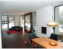 Modernes Wohnzimmer mit großen Fenstern, die einen schneebedeckten Außenbereich zeigen. Der Raum hat dunklen Holzboden, eine schwarze Eckcouch auf einem roten Teppich, einen Esstisch mit einer goldenen Hängelampe darüber und einen Kamin in der weißen Wand.