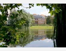 Ein Backsteingebäude mit großen Fenstern, umgeben von Bäumen, spiegelt sich in einem ruhigen See wider. Der Vordergrund ist von Zweigen mit grünem Laub eingerahmt.