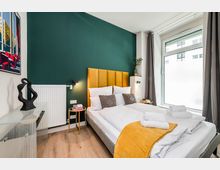 Modernes Schlafzimmer mit zwei Einzelbetten, senfgelbem gepolstertem Kopfteil vor dunkelgrüner Akzentwand und Holzfußboden. Rechts ein großes Fenster mit grauen Vorhängen; daneben kleine Nachttische mit Lampen, links eine Kommode und ein Wandbild.