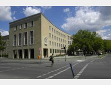 Eckgebäude in heller Steinoptik mit großen, rechteckigen Fenstern und einem Relief an der Fassade. Vor dem Gebäude befindet sich ein Bürgersteig, ein Fußgängerüberweg und einige Bäume; im Vordergrund läuft eine Person über die Straße.