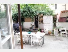 Hinterhof mit zwei Wohnwagen, umgeben von dichtem Efeubewuchs und Pflanzen. Im Vordergrund steht ein weißer Klapptisch mit Stühlen auf einem gepflasterten Boden; daneben liegen Gegenstände wie Blumentöpfe und Fensterrahmen.