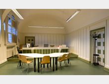 Ein heller Raum mit gewölbter Decke, einer Empfangstheke im hinteren Bereich und einem weißen Tisch mit vier Holzstühlen im Vordergrund. Die Wände sind paneelverkleidet, links befinden sich große Fenster, rechts ein Kleiderständer, und die Decke ist mit Hängelampen ausgestattet.