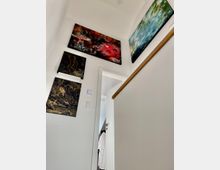 Treppenaufgang in einem modernen Innenraum mit weißen Wänden, an denen abstrakte Kunstwerke in kräftigen Farben hängen, und einem Holzhandlauf rechts. Im Hintergrund ist eine geöffnete Tür mit einem Einblick in einen weiteren Raum sichtbar.