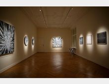 Ein heller Galerieraum mit Holzboden und Stuckdecke, in dem abstrakte, geometrische Kunstwerke an den Wänden ausgestellt sind, darunter runde und quadratische Gemälde mit strahlenden Mustern in Schwarz-Weiß. Rechts steht ein kleiner Klapptisch vor großen Fenstern.