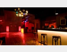 Eine stilvolle Lounge mit Holzboden, einer langen beleuchteten Bar und Hockern im Vordergrund. Im Hintergrund befindet sich ein dekorativer Bereich mit hängenden orientalischen Lampen, roten Lichtakzenten und einem Vorhang aus Ketten.