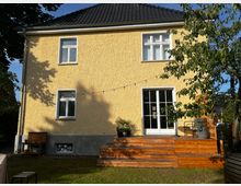 Zweistöckiges Wohnhaus mit gelber Putzfassade und schwarzen Ziegeldach. Im Vordergrund befindet sich eine hölzerne Terrassenkonstruktion mit Blumenkübeln und Sitzmöbeln, umgeben von einem Garten mit Rasen und Bäumen.