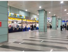 Innenbereich eines Bahnhofs mit einem Supermarkt mit der Aufschrift „Edeka City-Markt“ auf der linken Seite. Der Bereich hat helle Deckenbeleuchtung, breite Fliesenböden mit Pfeilen, mehrere moderne Säulen und eine Treppe im Hintergrund.