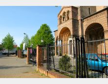 Backstein-Kirchgebäude mit rundbogigen Eingangspforten im romanischen Stil, flankiert von Säulen. Im Vordergrund ein schmiedeeiserner Zaun mit gemauerten Pfeilern und parkenden Autos, umgeben von Grünflächen und Bäumen.