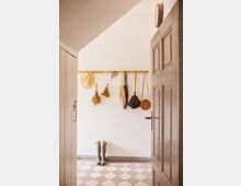 Blick in einen schlichten Eingangsbereich mit weiß gestrichenen Wänden und einem Boden in Schachbrettmuster. An einer Holzgarderobenleiste hängen Hüte, Taschen und getrocknete Blumen, und unten stehen Gummistiefel.