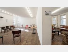 Offener Raum mit Holzboden und weißen Wänden, eingerichtet mit verschiedenartigen Stühlen, Tischen und Sesseln. Tageslicht fällt durch große Fenster, eine Leiter steht an einer Wand, und ein Schild weist auf "LAFT SUPERpolis" hin.