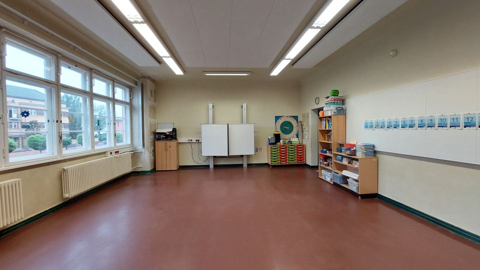 Klassenzimmer mit braunem Boden, großen Fenstern auf der linken Seite und weißen Wänden. Vorne befinden sich eine interaktive Tafel, Regale mit Materialien und ein Kalender an der Wand. Außerhalb der Fenster sind Schulgebäude und Grünflächen sichtbar.
