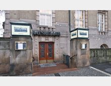 Eingang eines historischen Gebäudes mit der Beschriftung "Ratskeller" über Holztüren, flankiert von beleuchteten Schildern mit der Aufschrift "Ratskeller Restaurant". Die Fassade besteht aus Stein, und es sind hohe Fenster sichtbar.