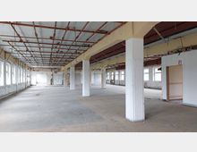 Großer, leerer Innenraum eines Gebäudes im Rohbauzustand mit freiliegender Decke, sichtbarer Metallkonstruktion und mehreren Säulen. Auf einer Seite befinden sich viele Reihen von Fenstern, während der Boden und die Wände unfertig wirken.